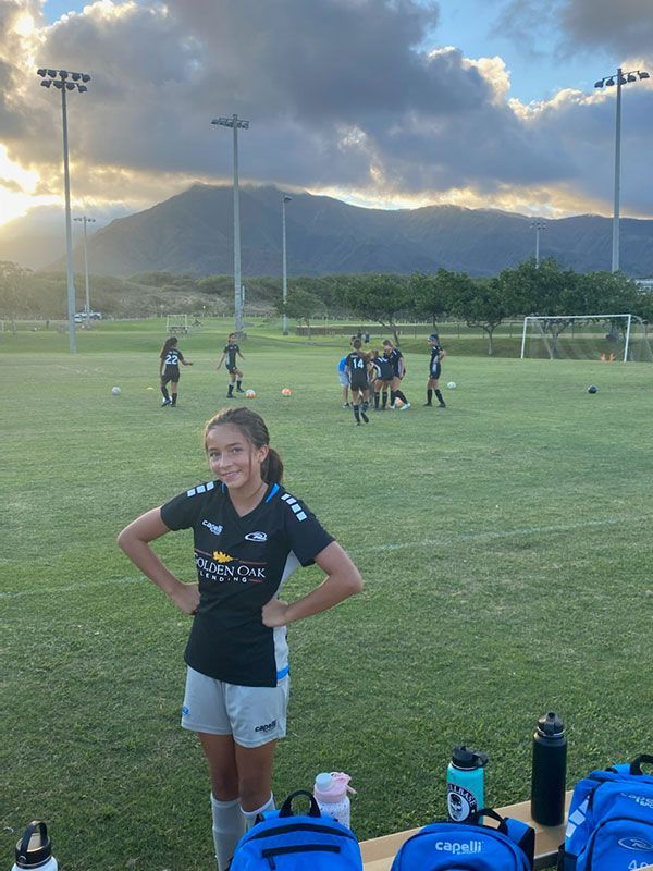 Young soccer player stands on field with hands on hips, other players in background. Mountains visible.