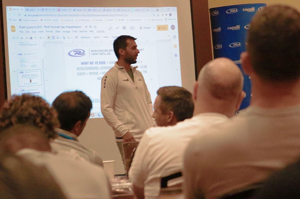 Man giving presentation to a seated audience, projected screen with text in the background.