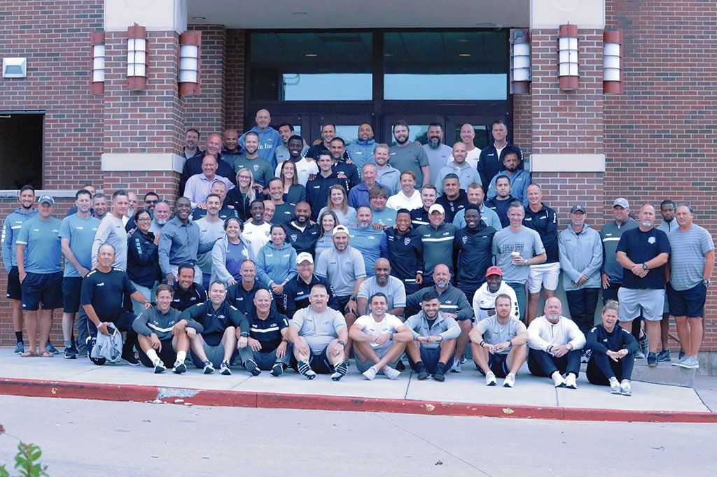 Group of people posing for photo in front of a brick building with a few people kneeling in the front.
