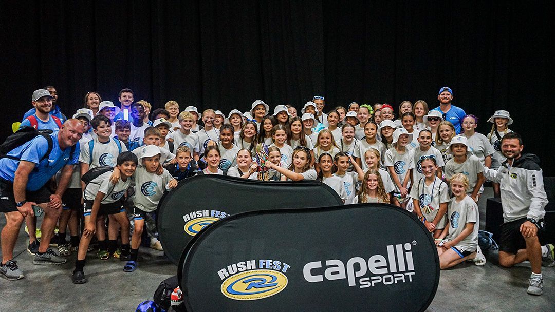 Large group of people in white and blue shirts, gathered for a photo. Black background. Capelli Sport banner in front.