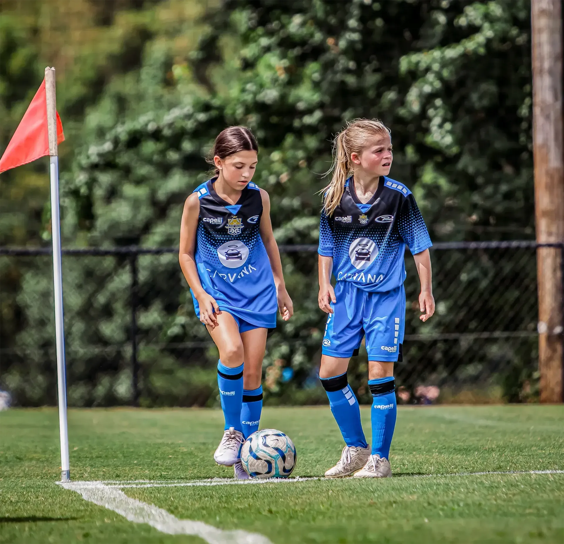 Two girls in blue soccer uniforms near a corner flag on a green field. One is about to kick the ball.
