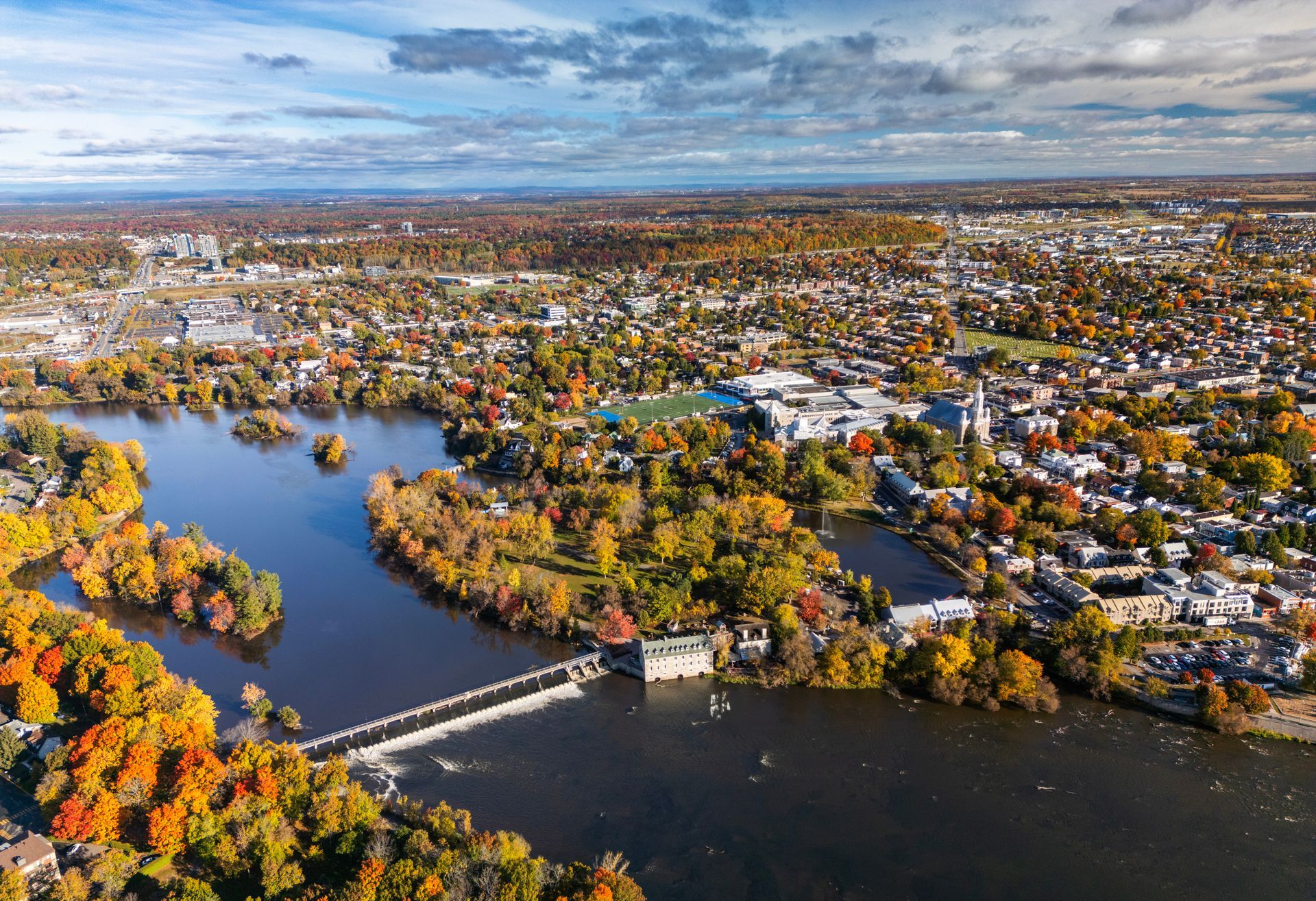 Vue aérienne d'une rivière avec des îles, un pont et des bâtiments, entourée d'arbres d'automne aux couleurs vibrantes sous un ciel bleu.
