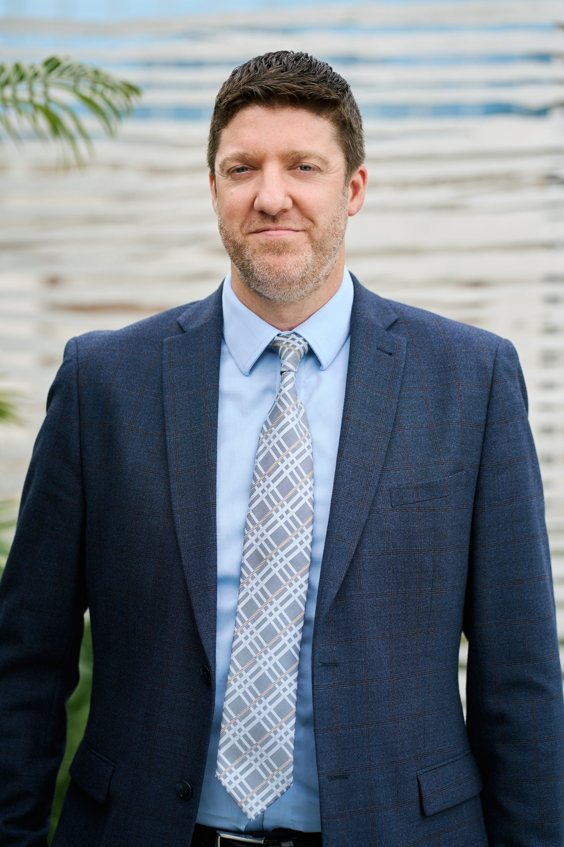 Un homme en blazer bleu marine et cravate sourit, debout devant un fond blanc et bleu.