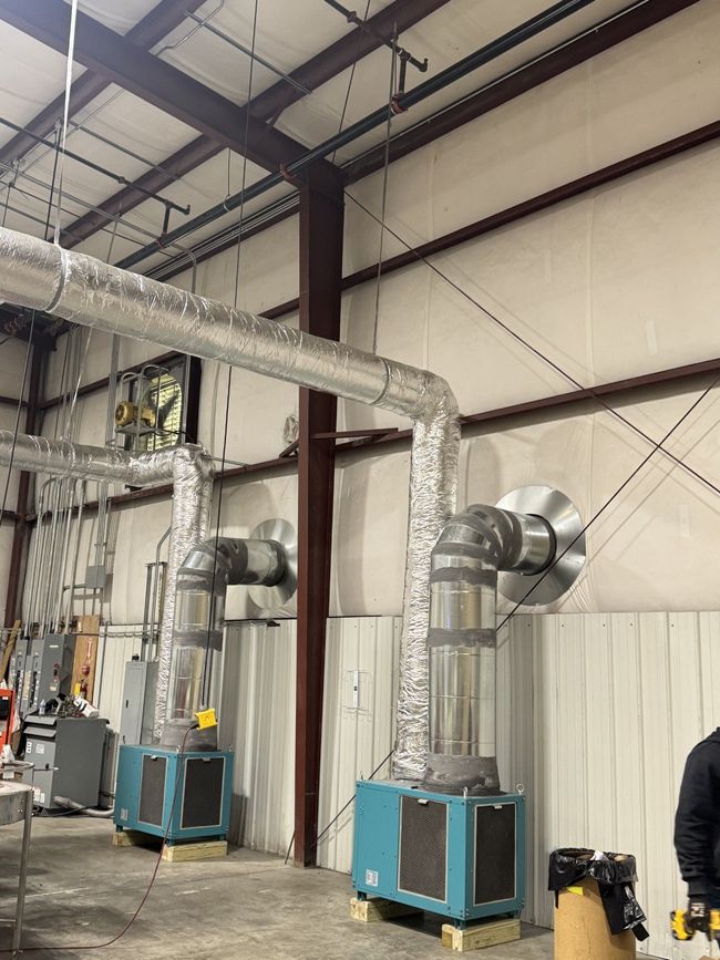 Industrial workshop with two blue portable HVAC units and silver ducting against a white metal wall.