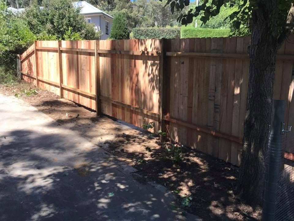 A Wooden Fence is Sitting on the Side of a Road Next to a Tree — A&H Fences In Moss Vale, NSW