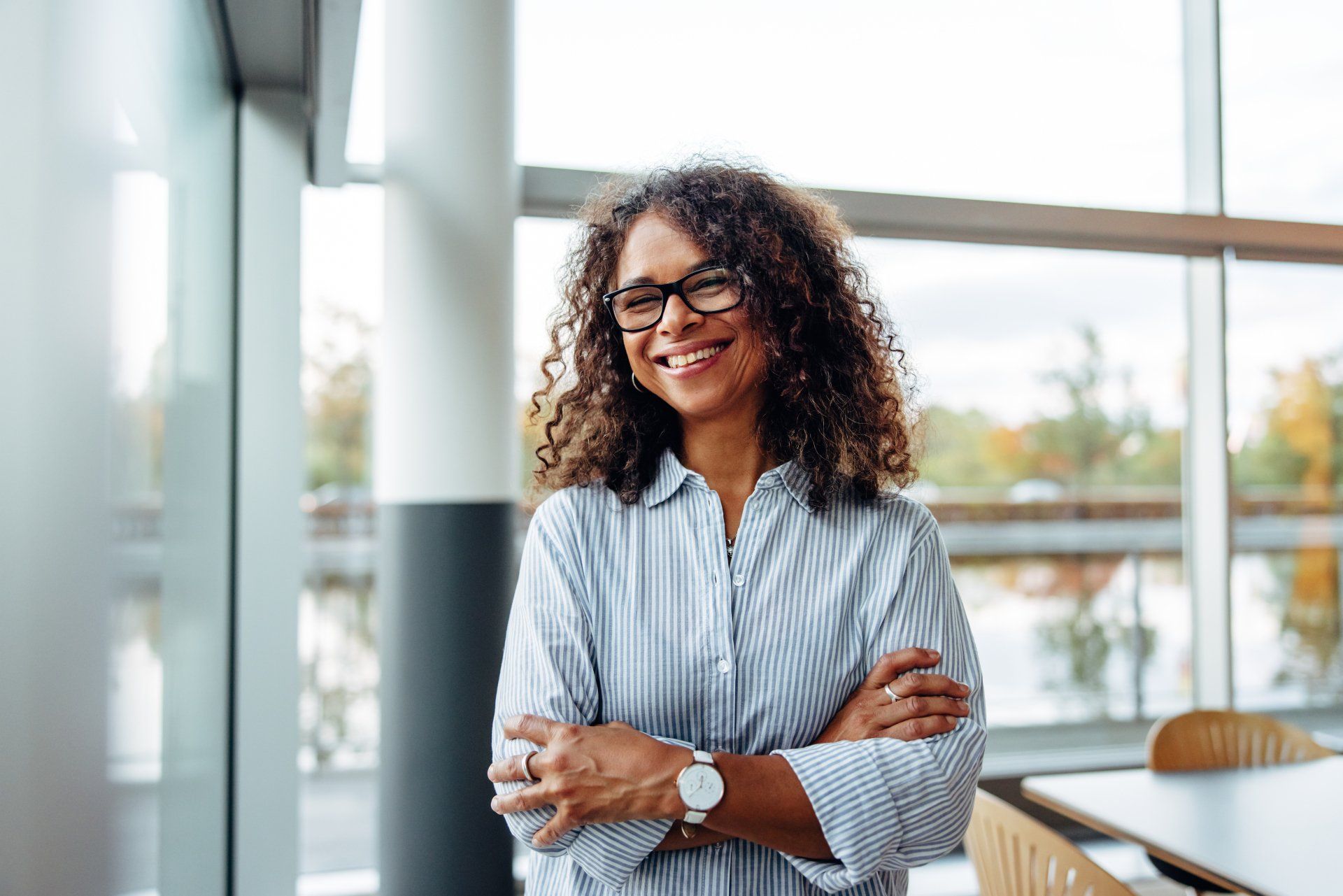 A woman with curly hair and glasses is standing in front of a window with her arms crossed and smiling.