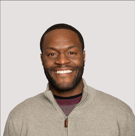 Smiling man with dark skin, wearing a gray zip-up sweater over a burgundy shirt.
