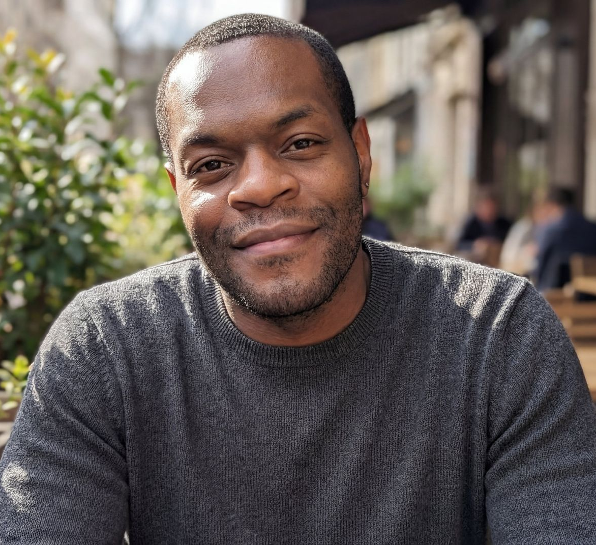 Smiling man with dark skin, wearing a gray zip-up sweater over a burgundy shirt.