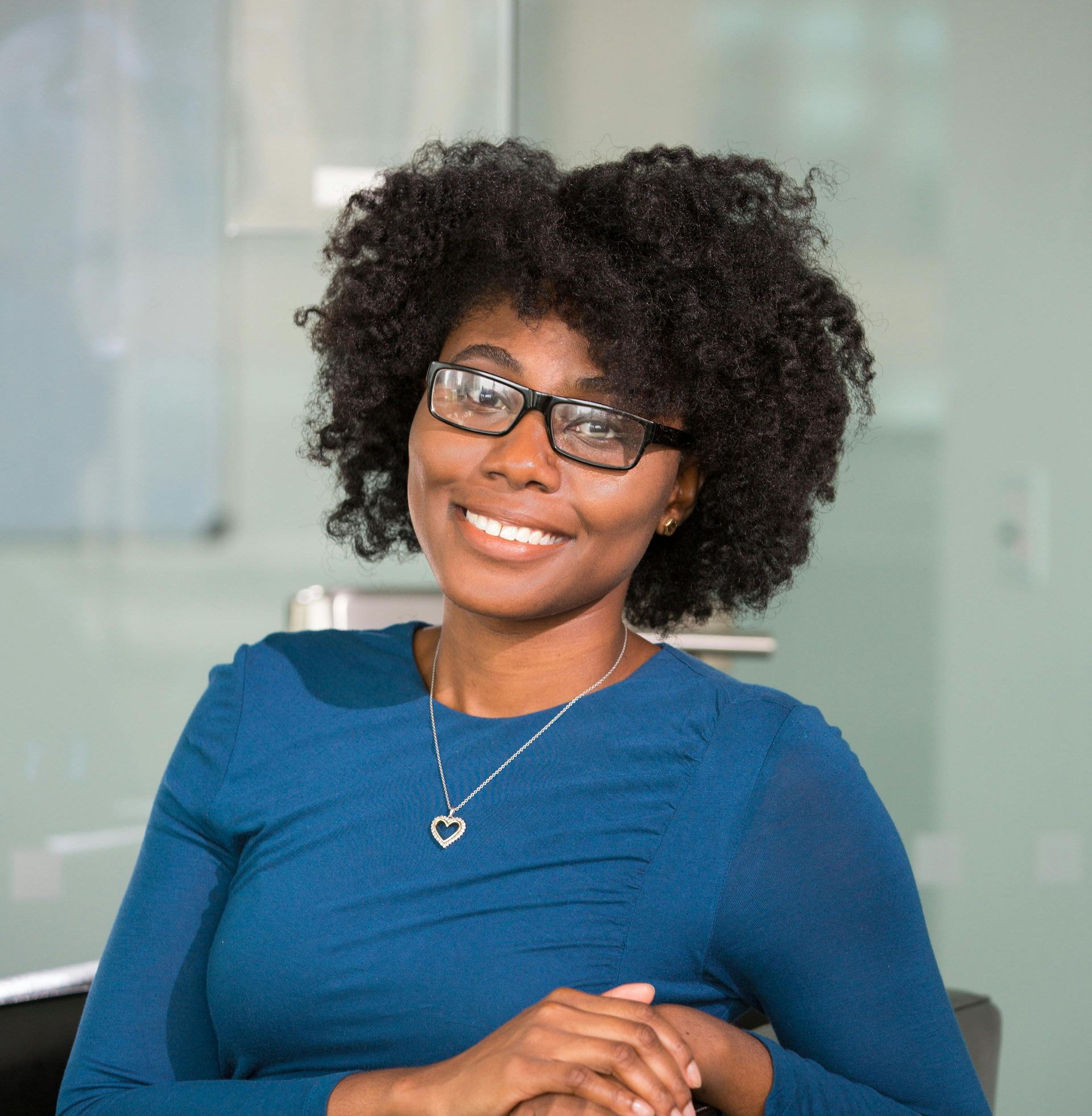 Woman with glasses smiles; wearing blue top and heart pendant, indoors.