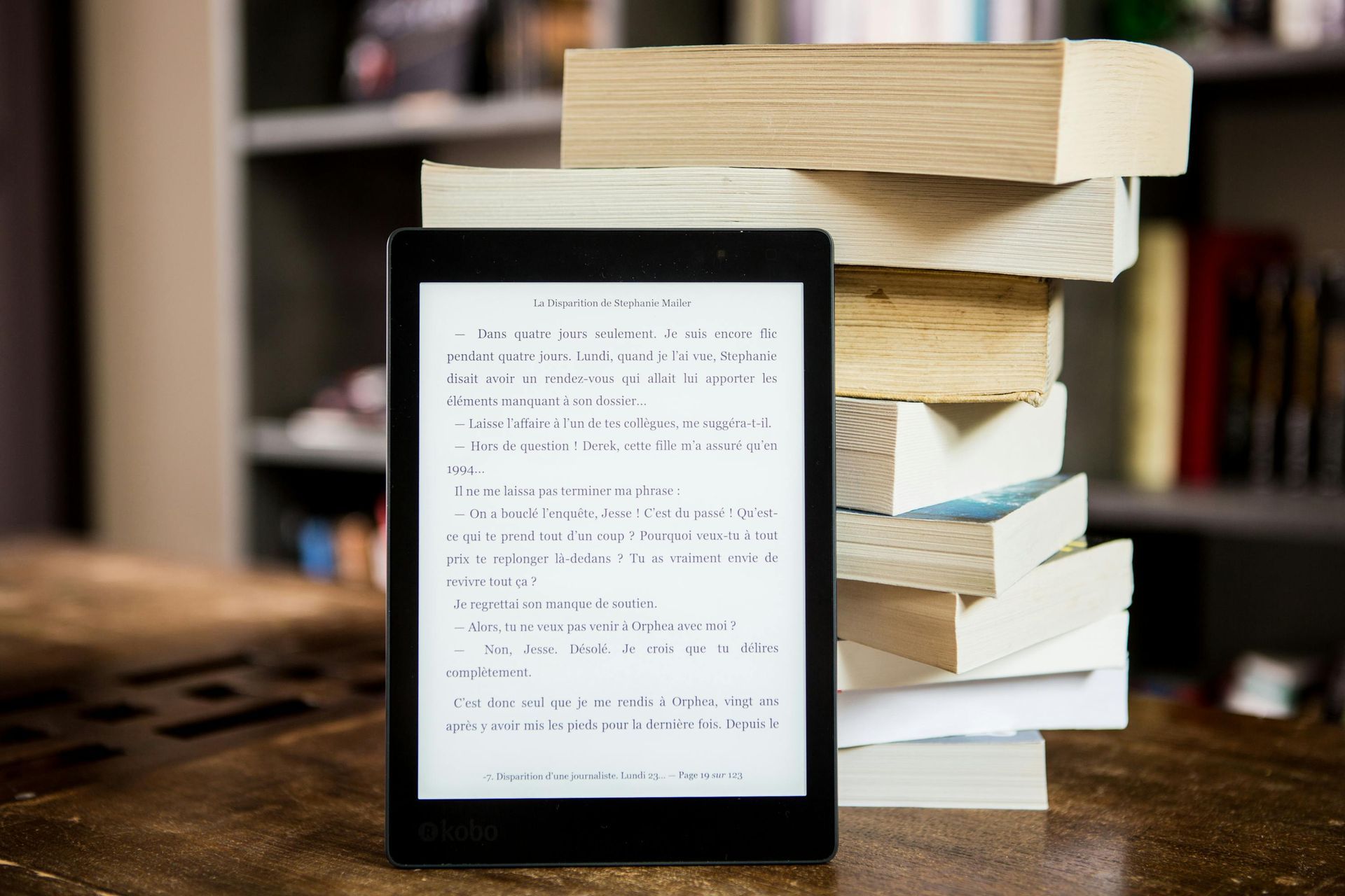 E-reader displaying text next to a stack of physical books on a wooden table; bookshelf in background.
