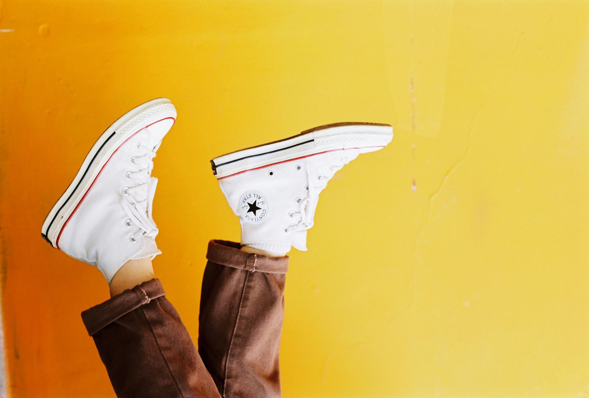 White high-top Converse sneakers on brown-clad legs, against a yellow background.