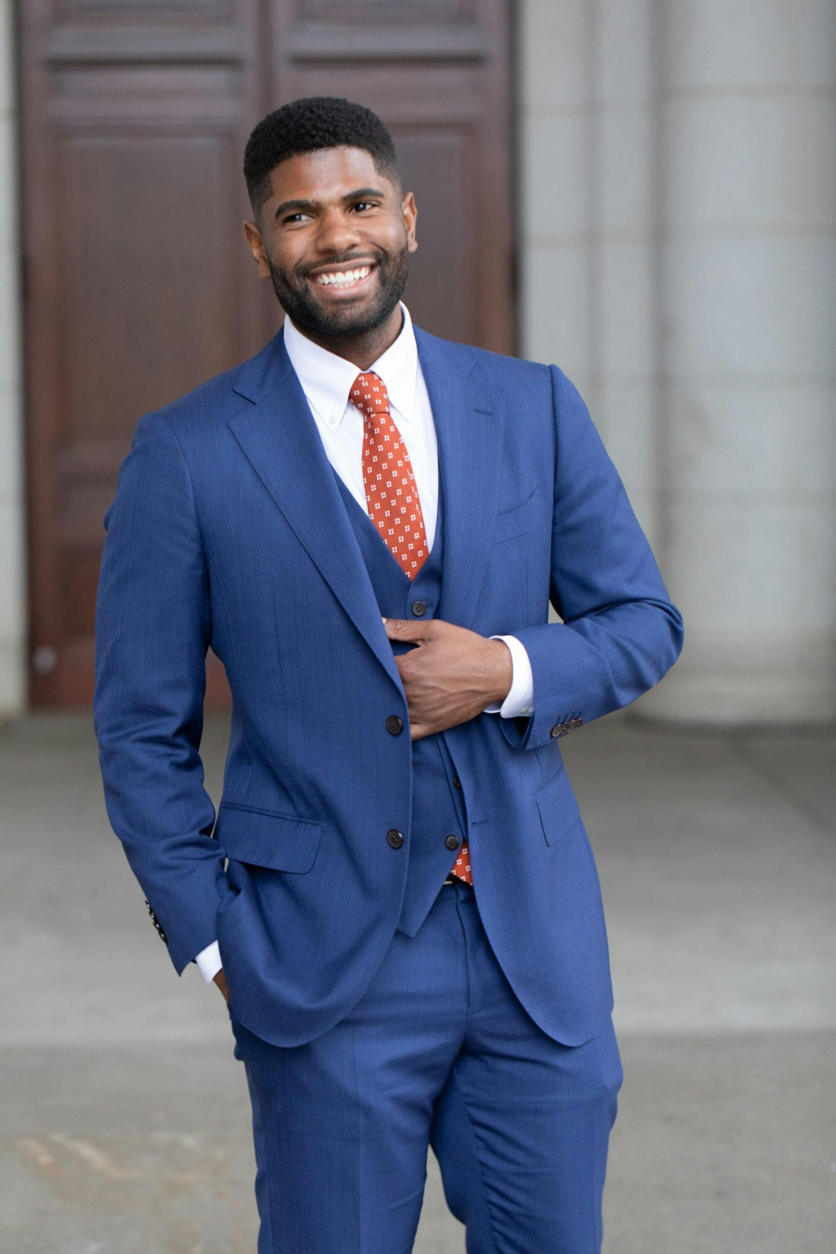 Man in blue suit, smiling, outdoors; holding suit jacket.