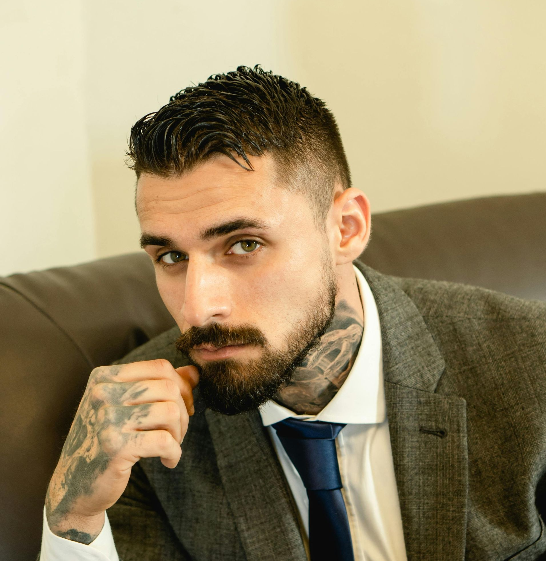 Man in gray suit and blue tie, hand on face, tattoos visible on neck and arm, sitting.