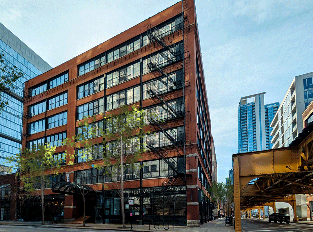 Industrial building with red brick and large iron windows in down town Chicago neighborhood. Elevated train sits right next to the building with cars driving by on a sunny day. 