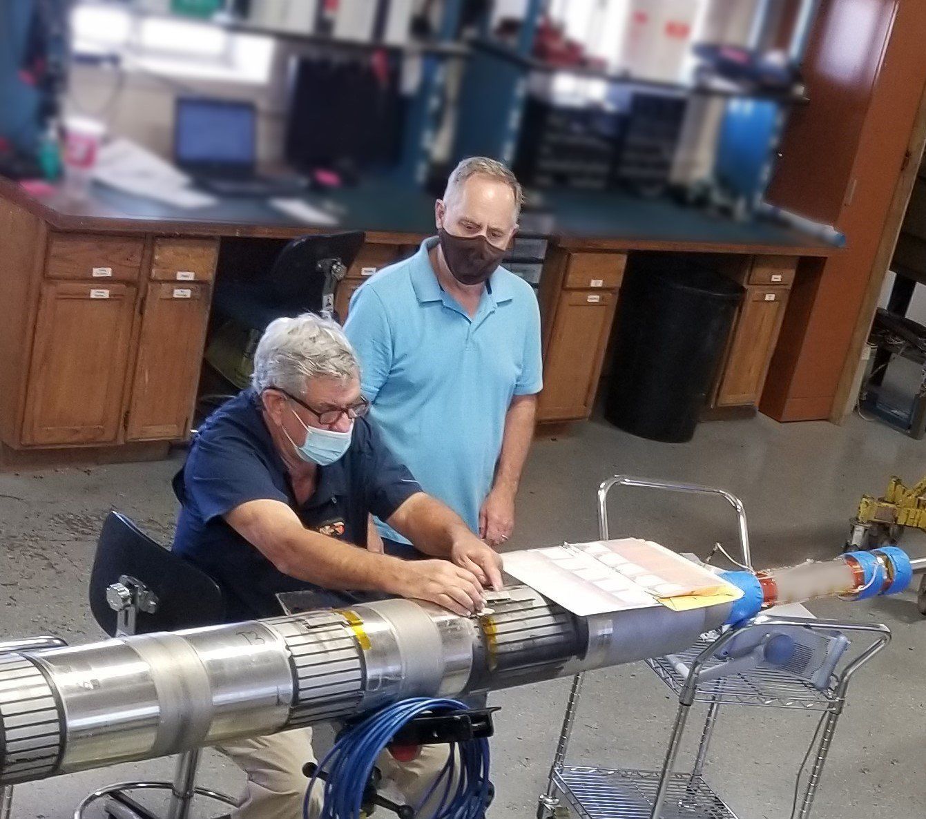 Two men wearing face masks are working on a large pipe.