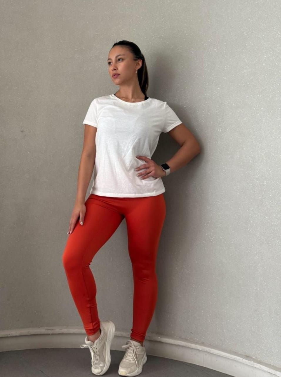 Mujer con camiseta blanca, leggings naranjas y zapatillas blancas, posa frente a una pared gris, con la mano en la cadera.
