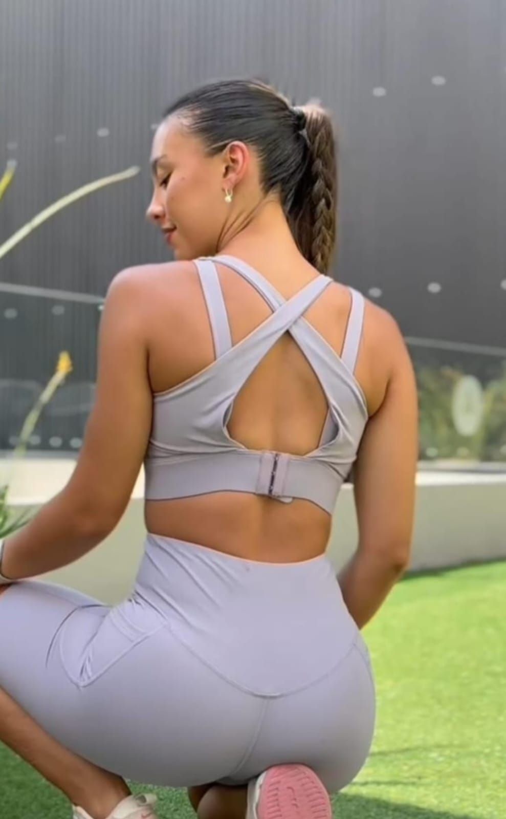 Mujer vestida con ropa deportiva gris, con cola de caballo, en cuclillas sobre el césped, mirando hacia abajo.