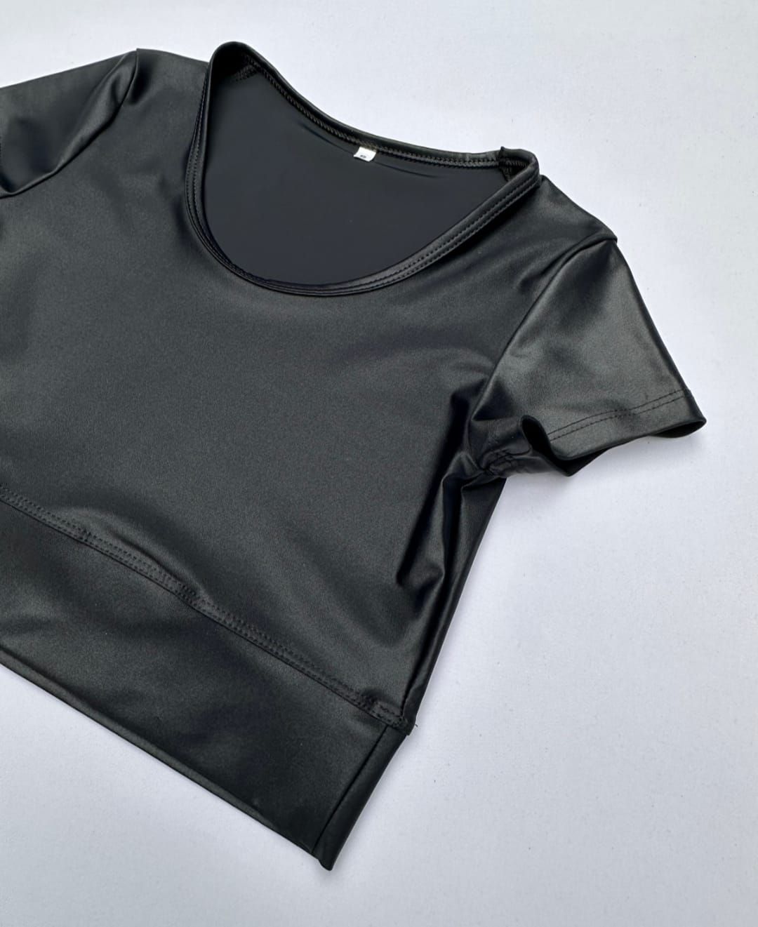 Top corto negro de manga corta y escote redondo. Tejido brillante sobre una superficie blanca.