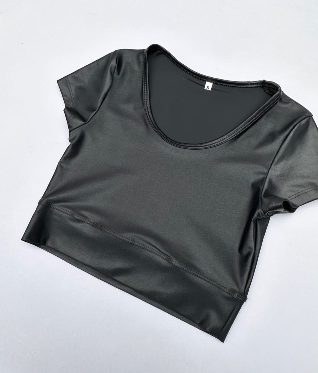 Top corto negro de manga corta y escote redondo. Tejido brillante sobre una superficie blanca.