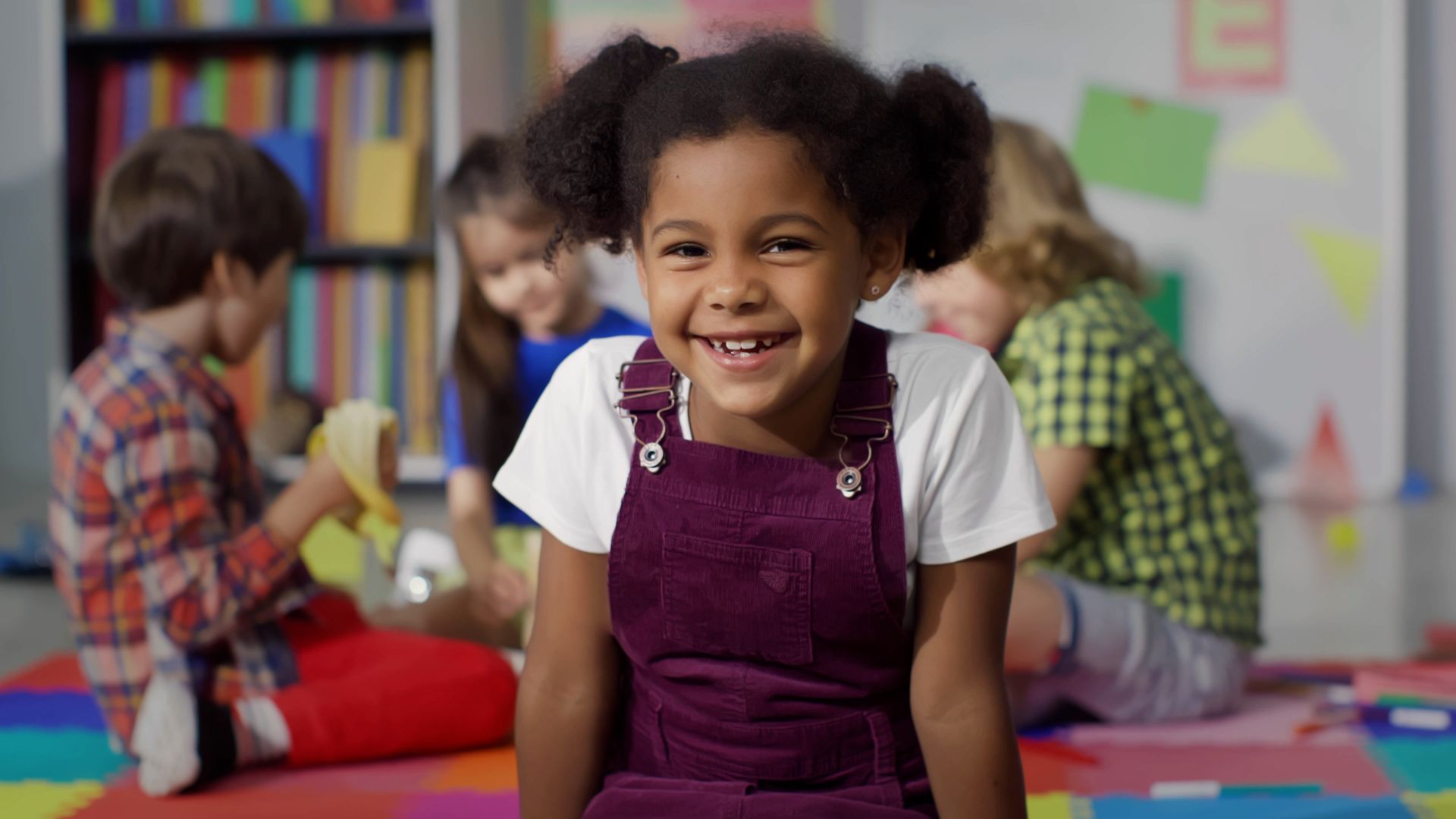 A little girl is sitting on the floor in a classroom with other children.
