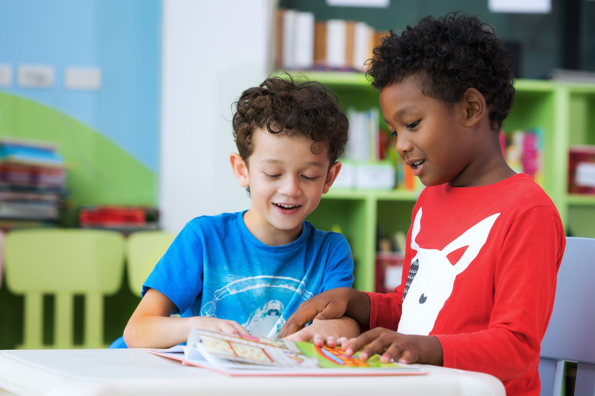 Two young boys are sitting at a table reading a book together.