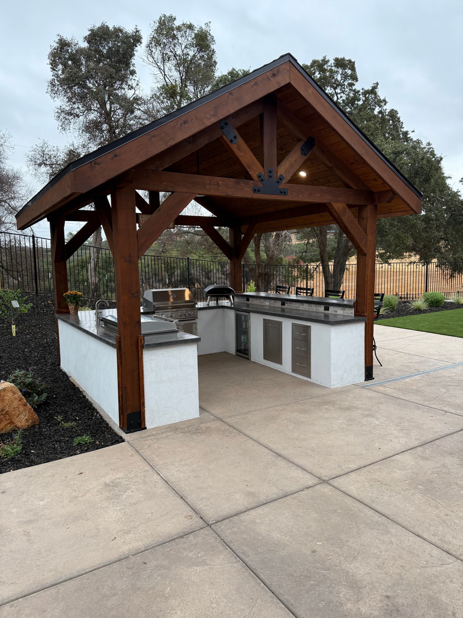 Outdoor kitchen under a brown wooden pergola with gray counters and stainless steel appliances.