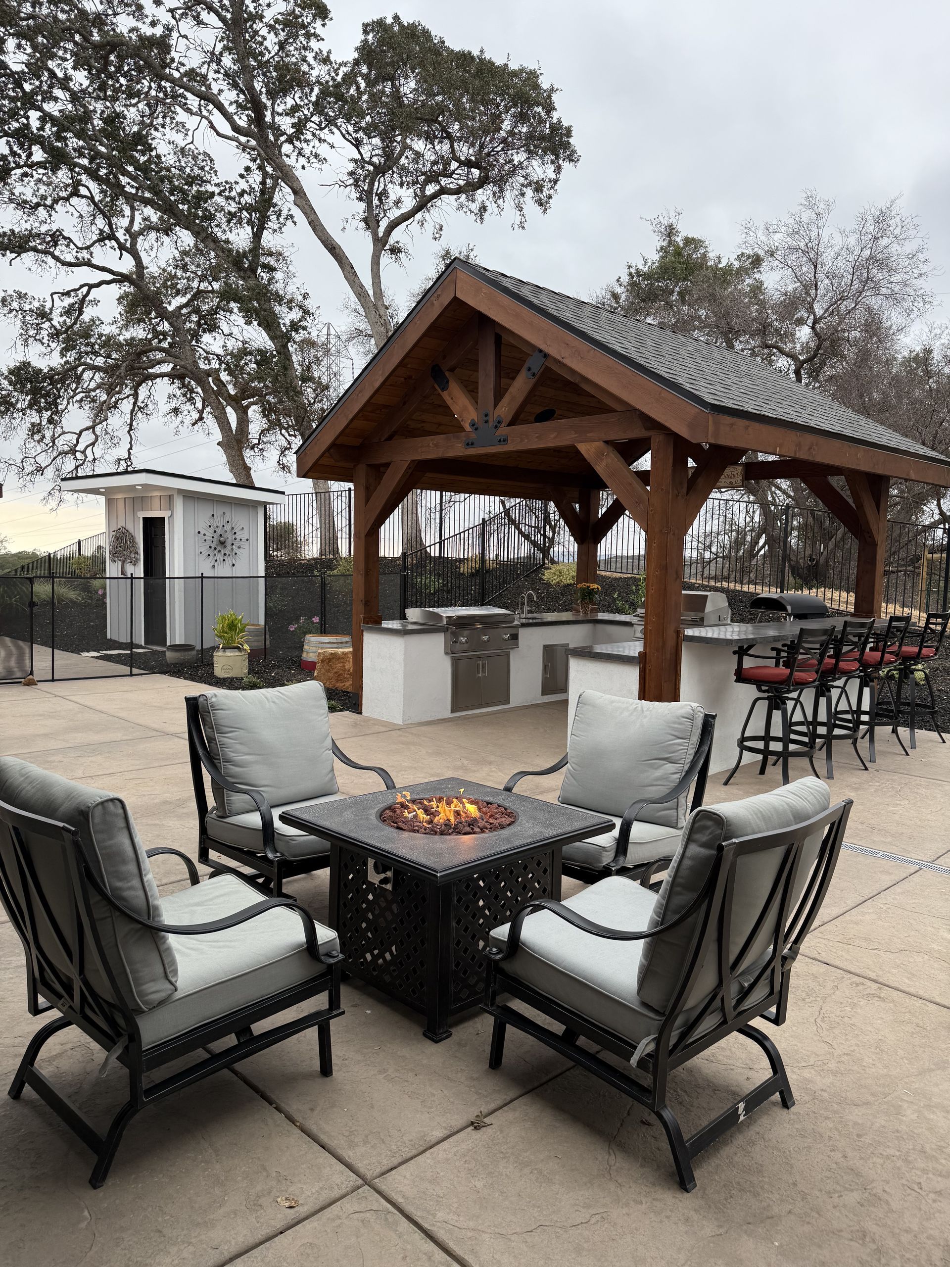 Outdoor kitchen and seating area with fire pit; dark wood pergola, gray cushions, overcast day.