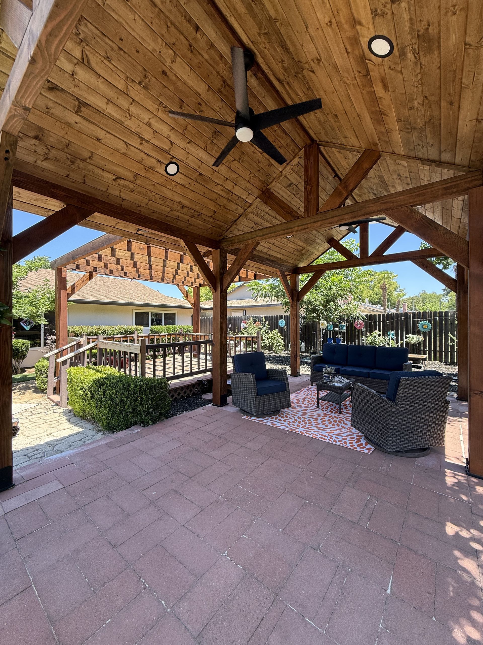 Wooden pavilion with wood beams, brick floor, and outdoor furniture; a ceiling fan hangs from the wood ceiling.