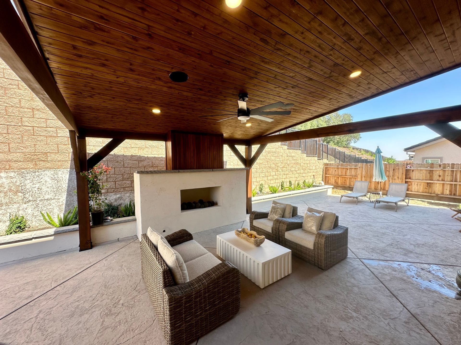 A patio with a fireplace , chairs , a table and a ceiling fan.