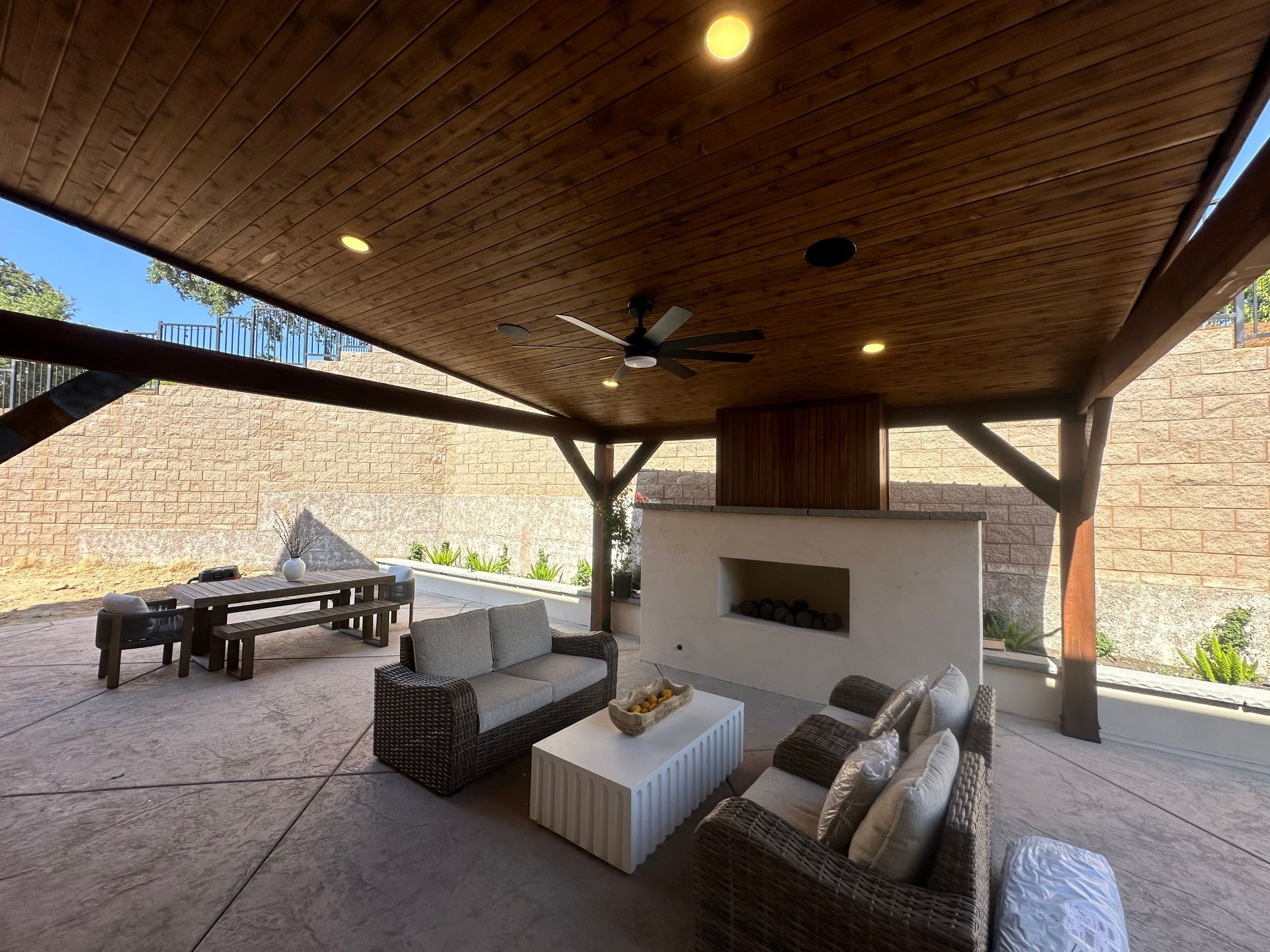 A patio with a couch , chairs , table and fireplace under a wooden roof.