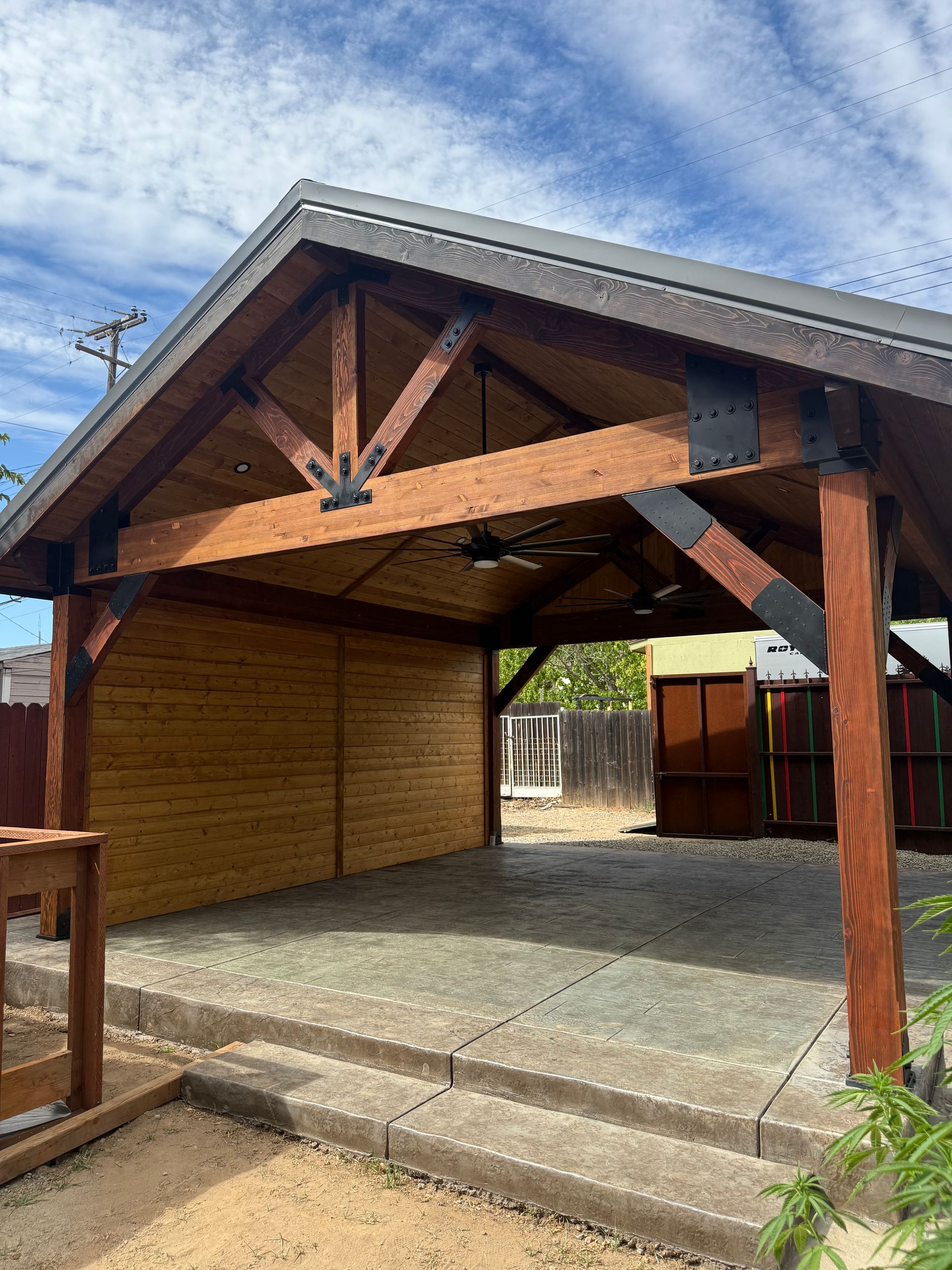 Wooden pavilion with brown beams, metal supports, and concrete patio under a blue sky.