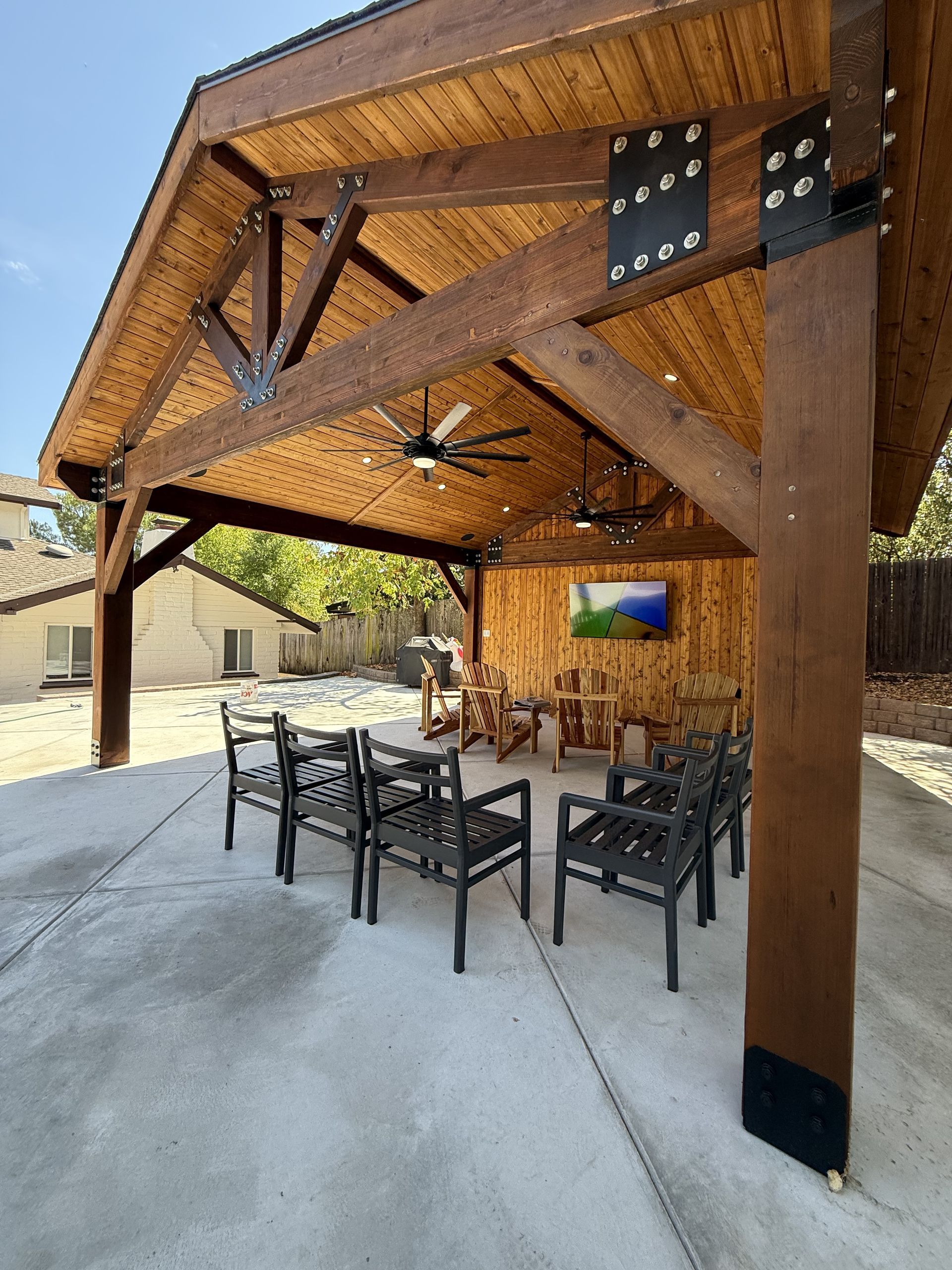 Wooden pavilion with seating area, fans, and TV, set on a concrete patio.