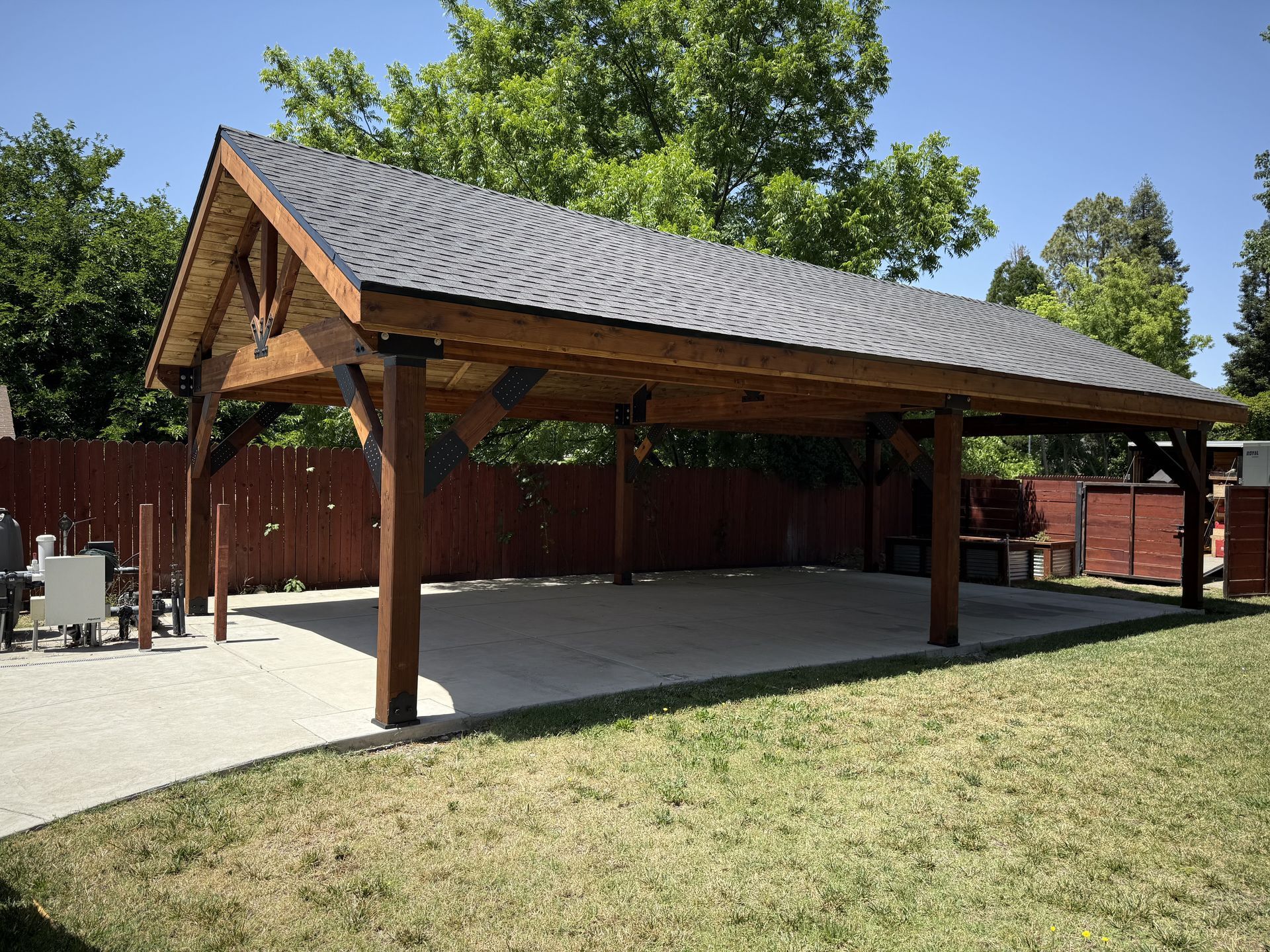 Wooden pavilion with dark roof on a concrete patio, red fence, and green lawn under a blue sky.