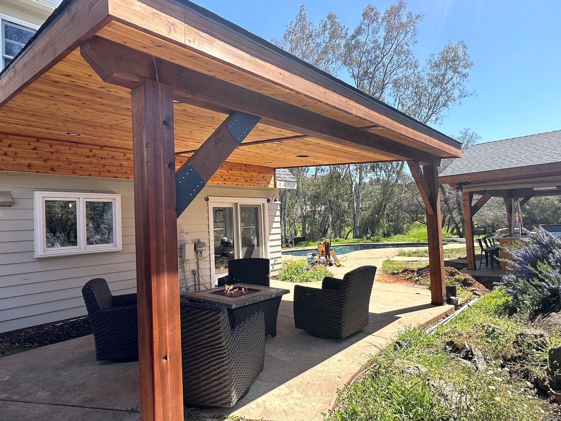 A patio with a fire pit and chairs under a wooden roof.