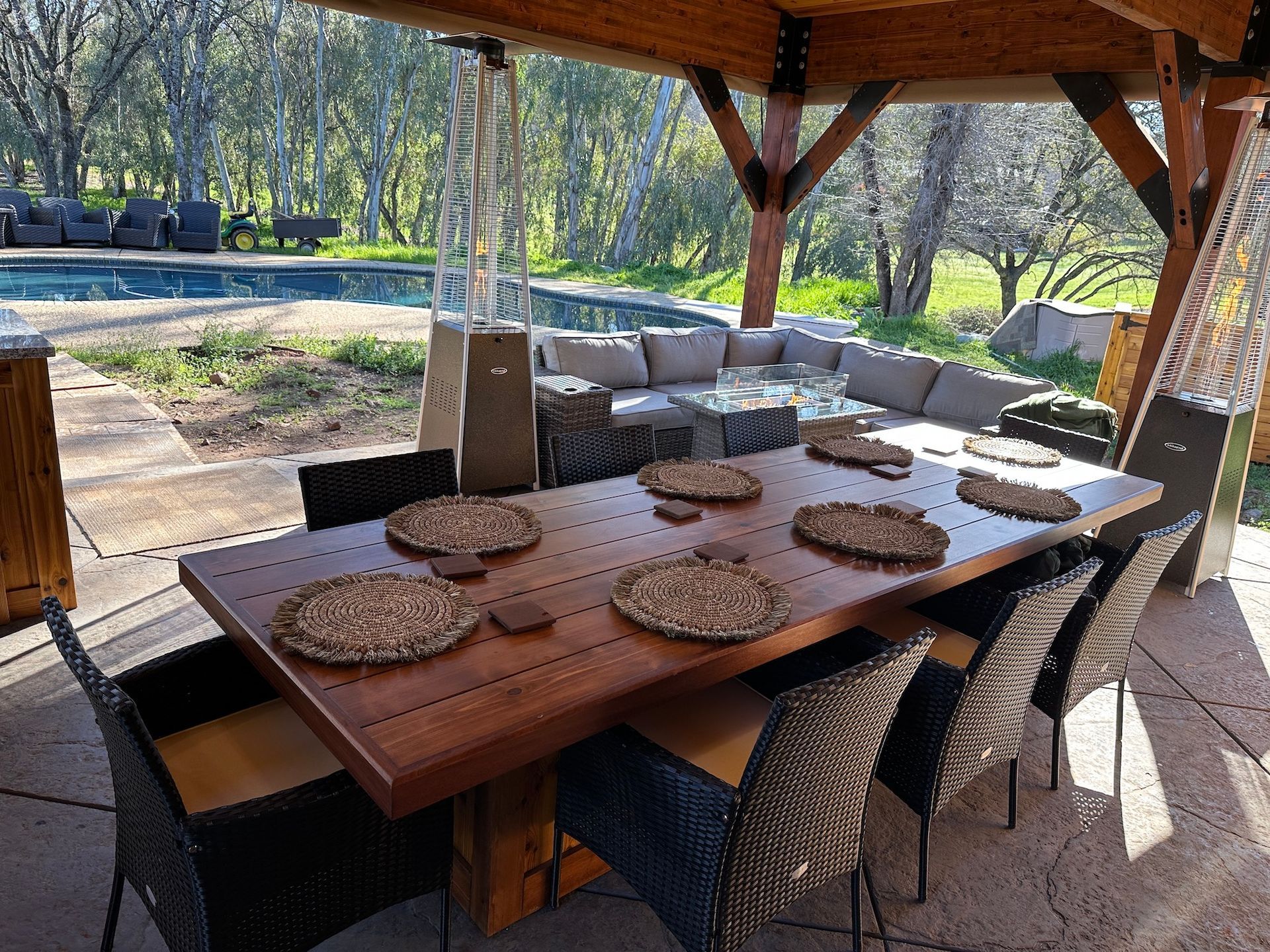 A large wooden table with wicker chairs under a gazebo with a pool in the background.