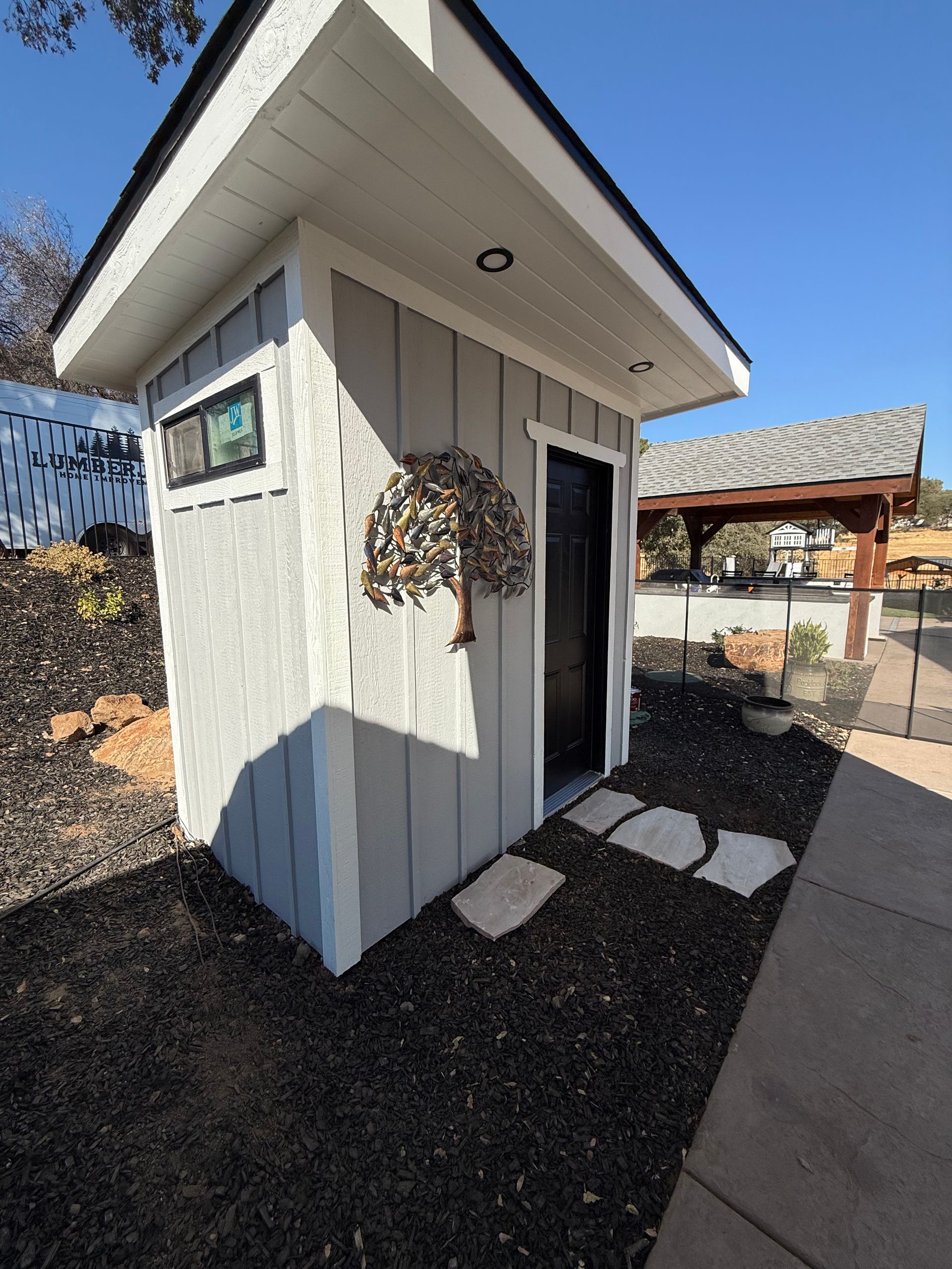 Gray shed with black door, stone path, and ornamental metal tree; sunny outdoors.