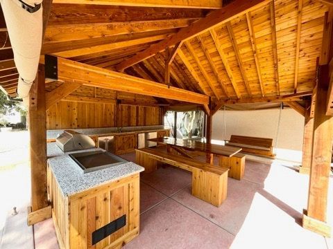 A wooden pavilion with a grill , table and benches under a wooden roof.