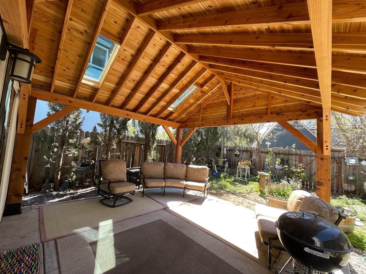 A wooden pavilion with a couch , chairs , and a grill.