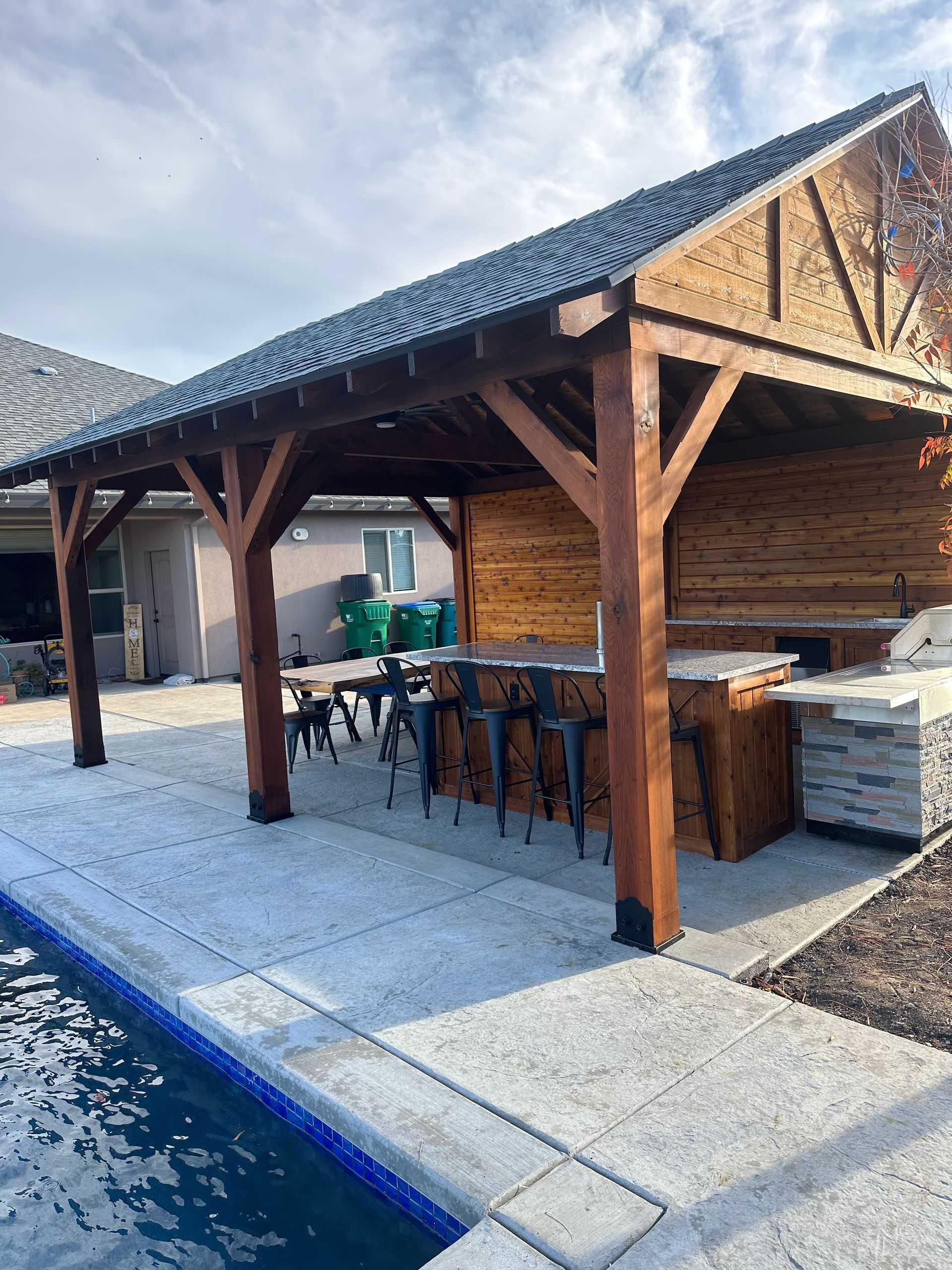 A wooden pavilion with a kitchen and a swimming pool in the background.