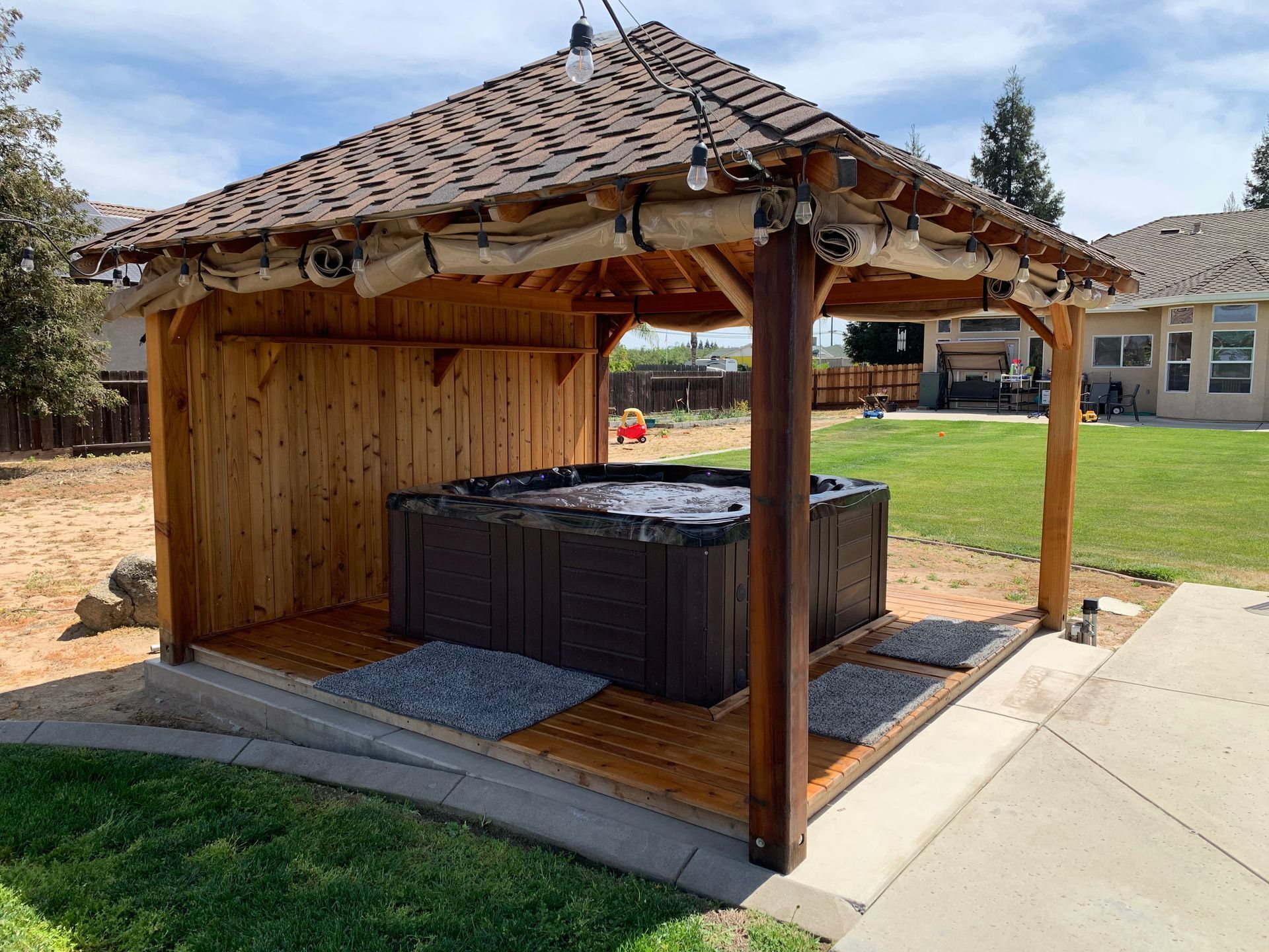 A hot tub is under a wooden pavilion in a backyard.