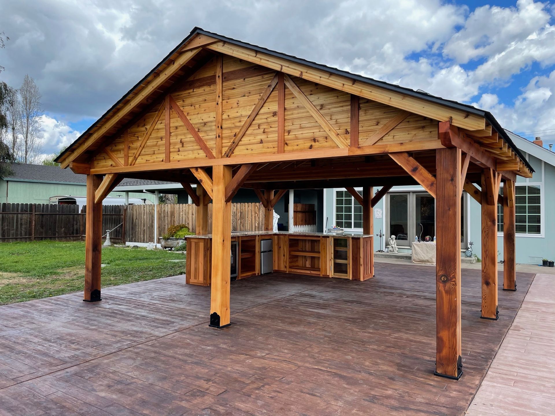 A large wooden pavilion with a kitchen underneath it is in the backyard of a house.