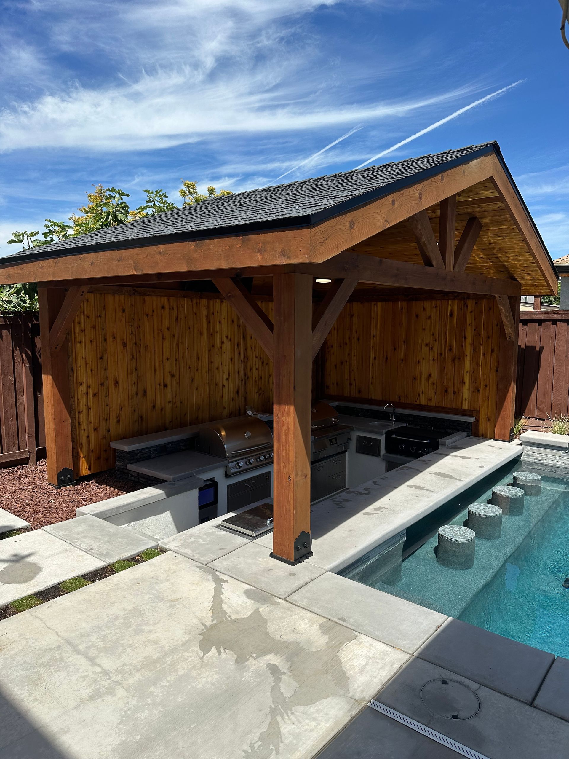 A wooden pavilion with a grill and a swimming pool in the background.
