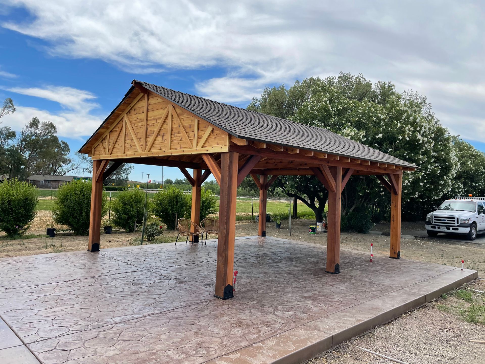 A car is parked under a wooden pavilion in a driveway.