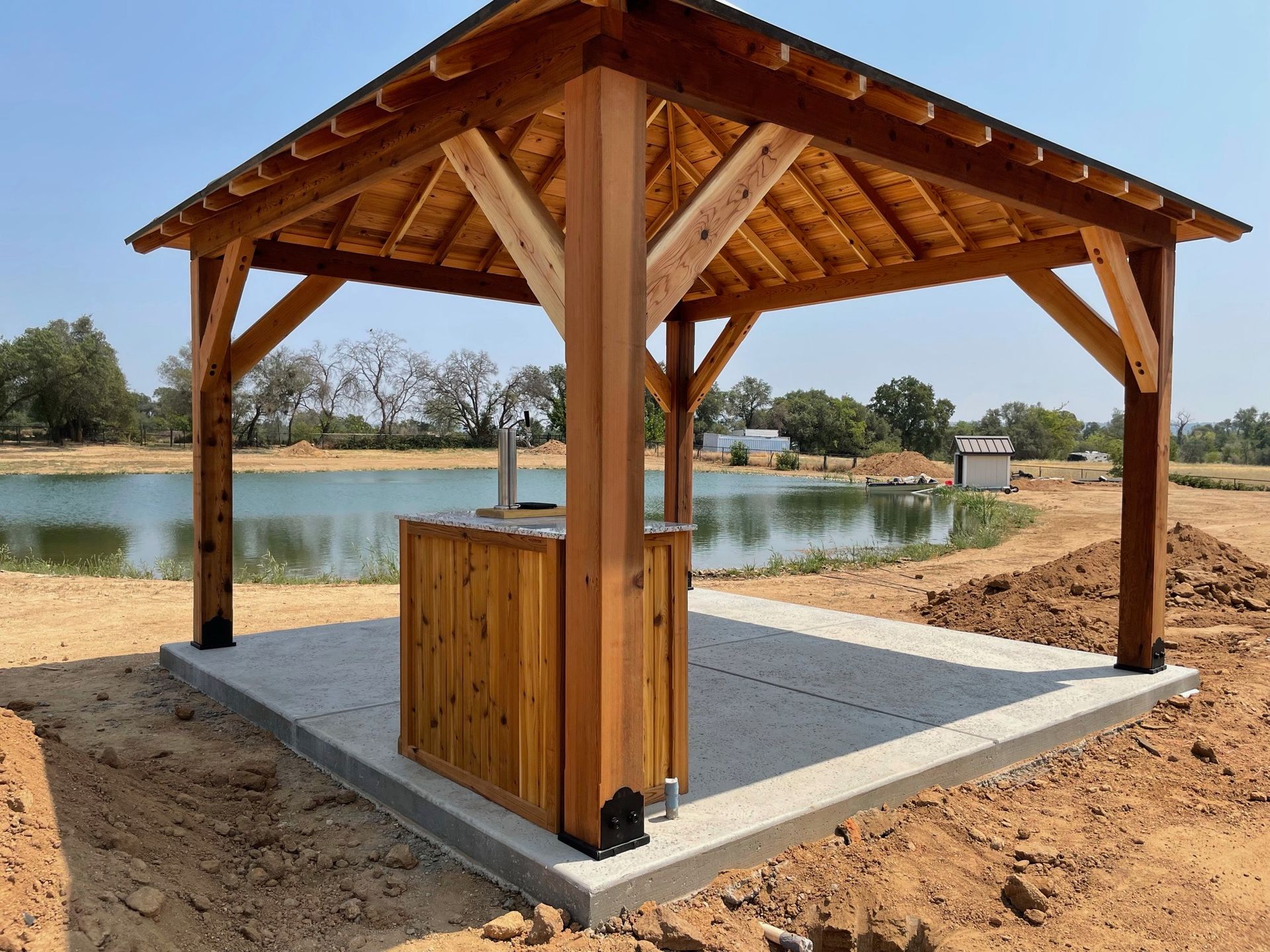 A wooden pavilion with a sink underneath it in front of a lake.