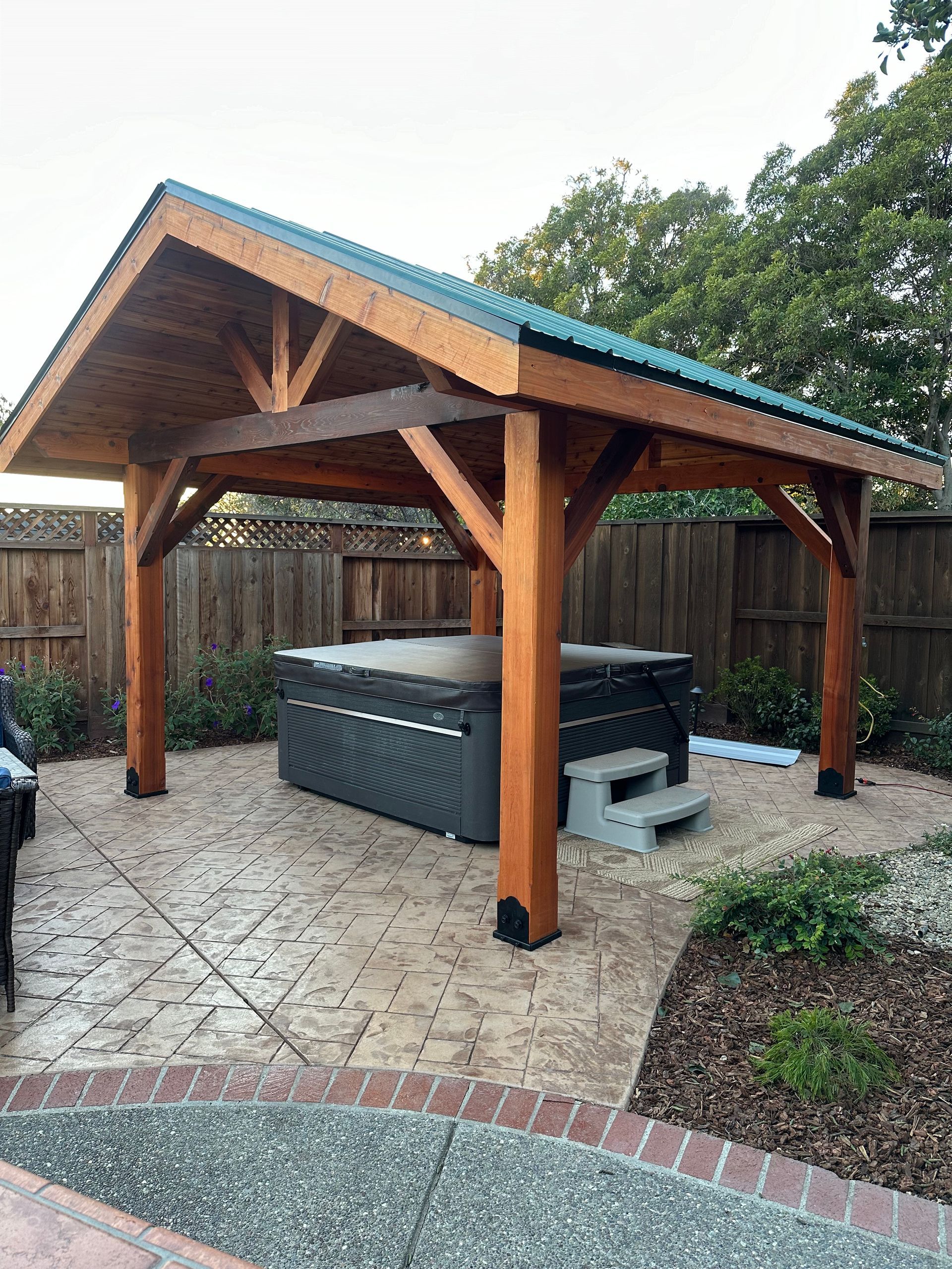 A hot tub is under a wooden pavilion in a backyard.