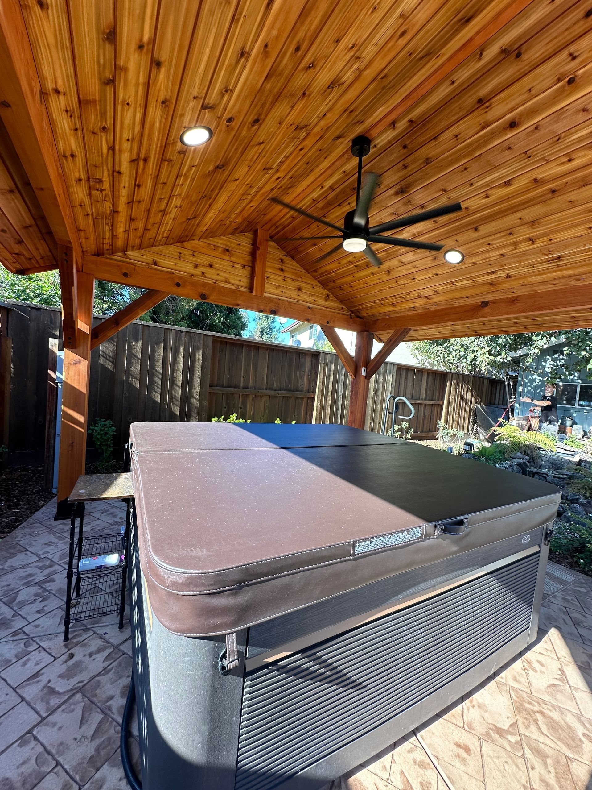 A hot tub under a wooden pavilion with a ceiling fan.