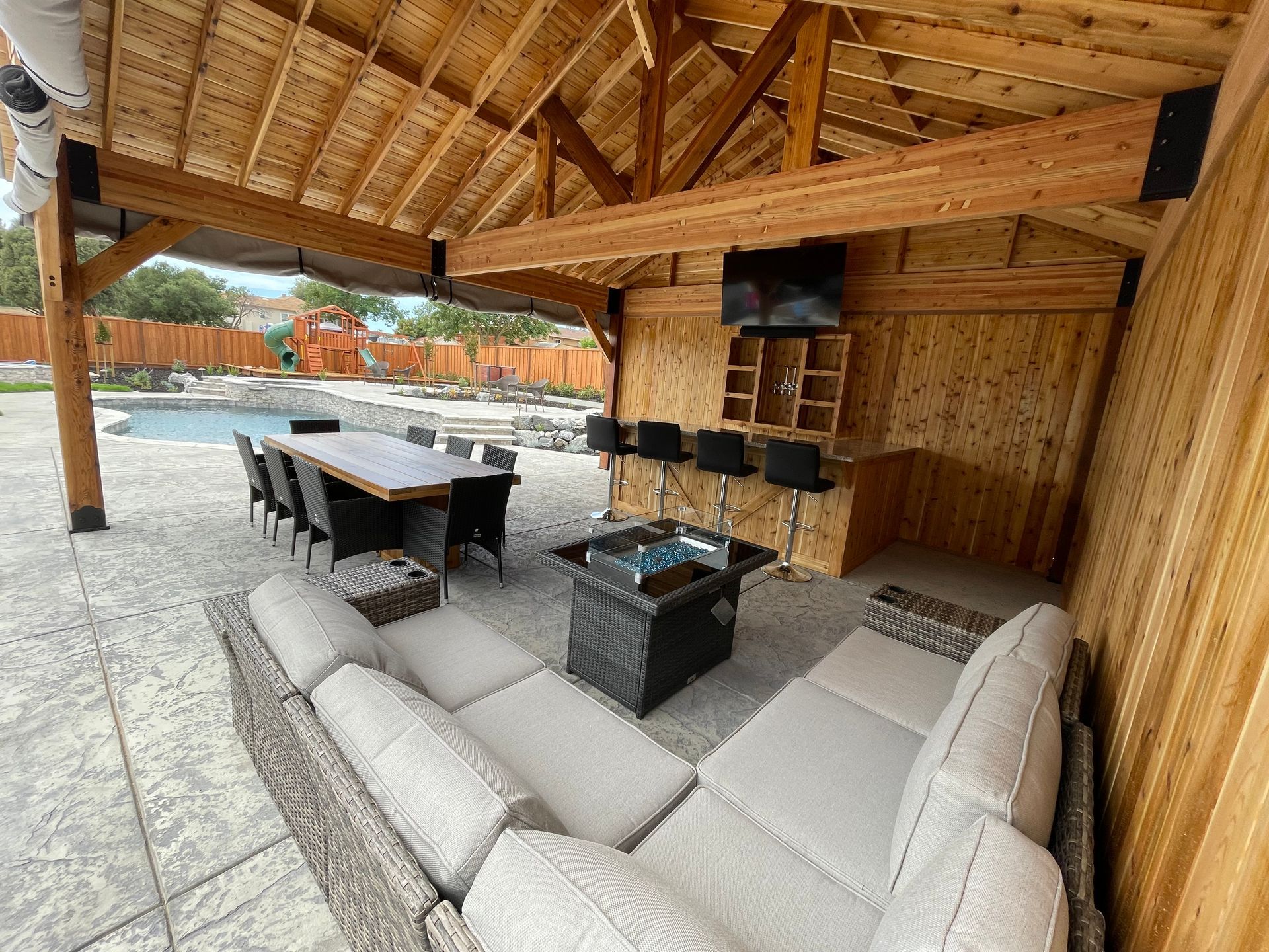 A patio with a couch , table and chairs under a wooden roof.