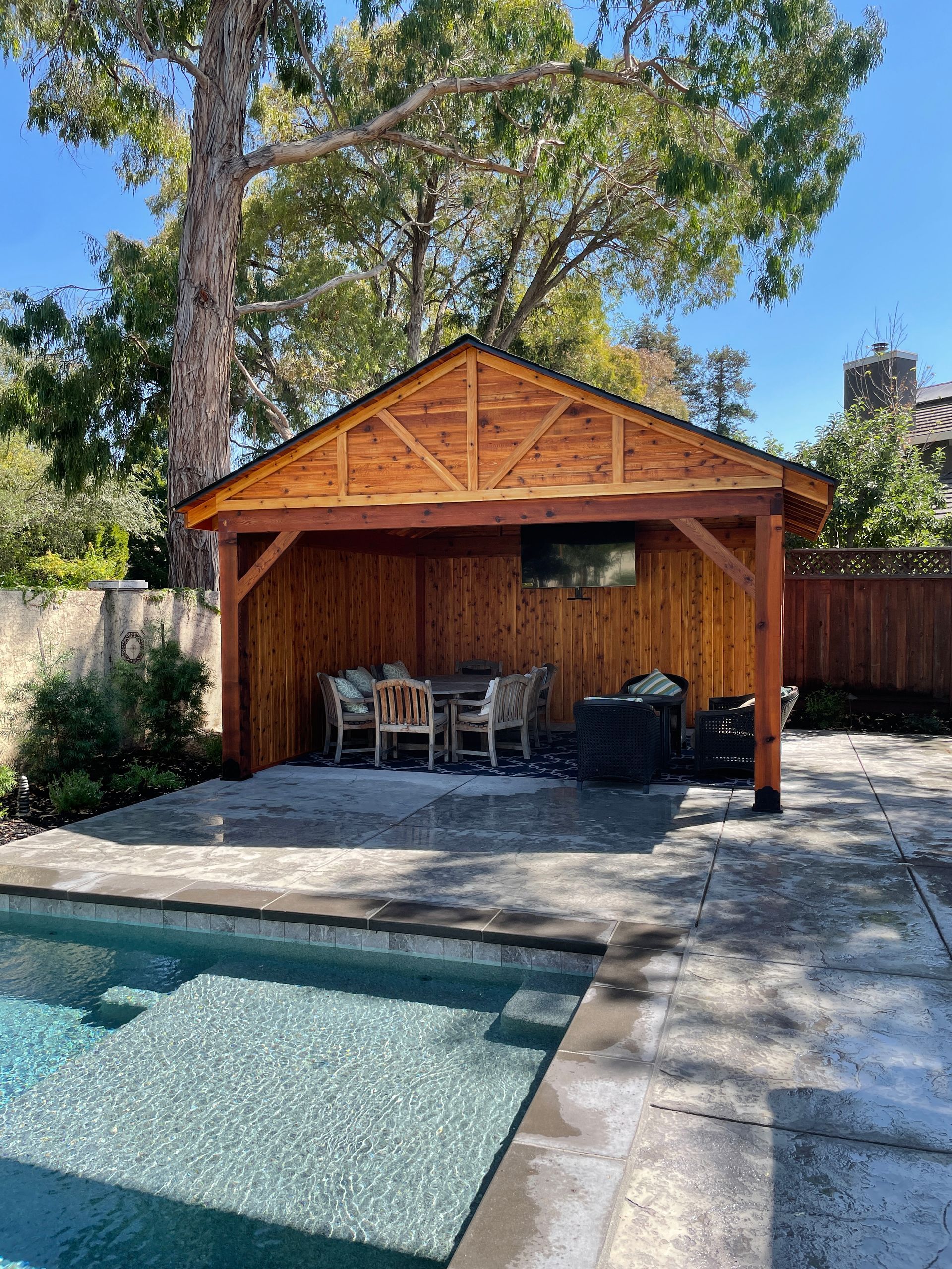 A wooden pavilion with a table and chairs next to a swimming pool.
