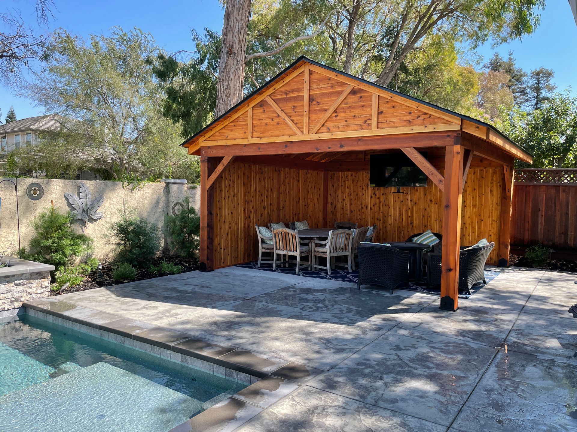 A wooden pavilion with a dining table and chairs next to a pool.