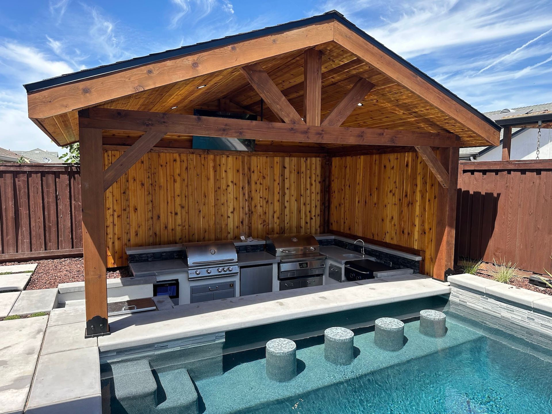 A wooden gazebo overlooking a swimming pool with a grill.